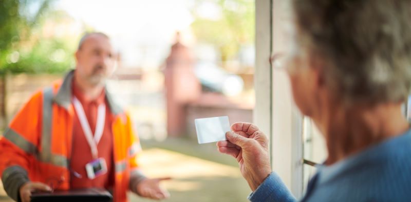 A photo of a man in a construction jacket at the door of a senior woman. She is looking at his business card.